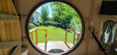 View through a circular window showing a green yard with trees and a wooden deck leading outside.